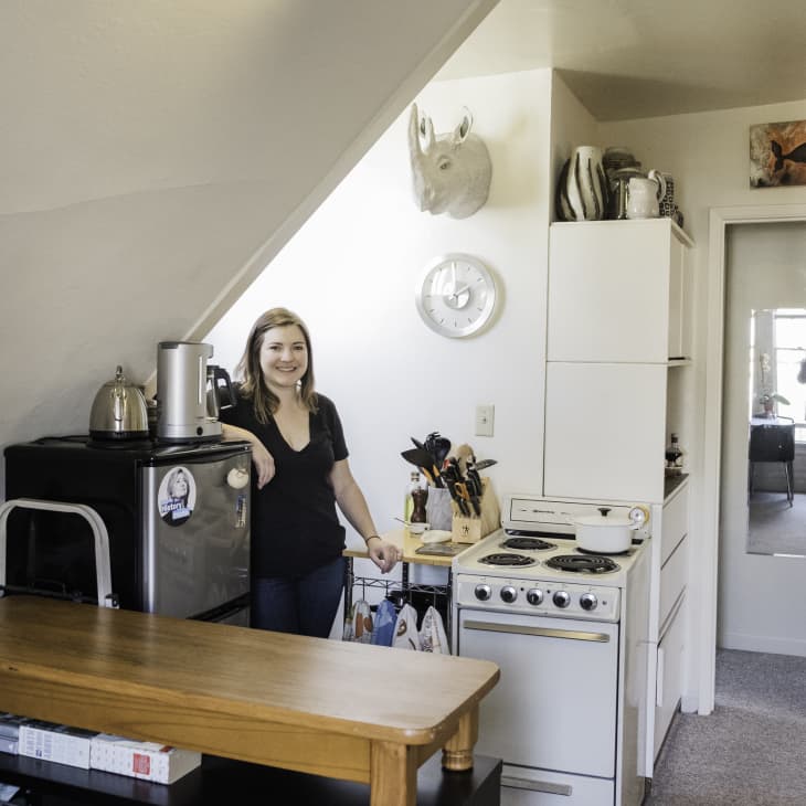 Woman in a small kitchen with a mini fridge, stove, and wooden counter, under a sloped ceiling with decor items.