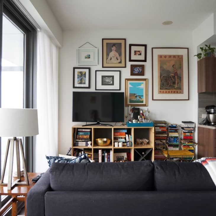 Living room with a dark sofa, wall-mounted TV, framed art, bookshelves, and a lamp by large windows.