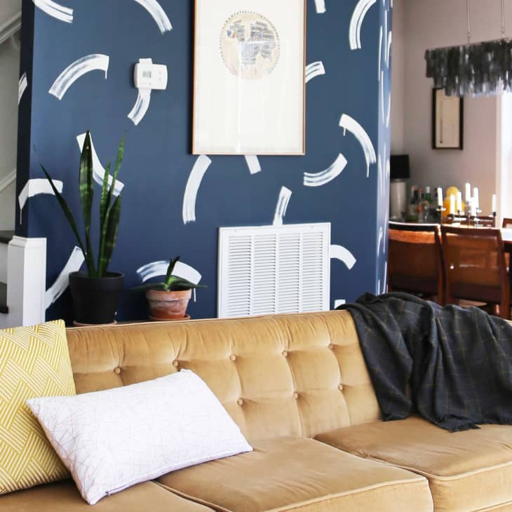 Tan sofa with yellow and white pillows, navy accent wall with abstract white patterns, and dining area in the background.