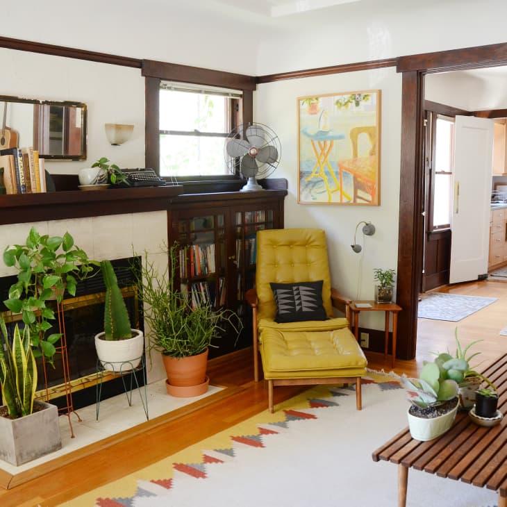 Living room with yellow chair, plants, wooden coffee table, and fireplace, leading to dining area and kitchen.
