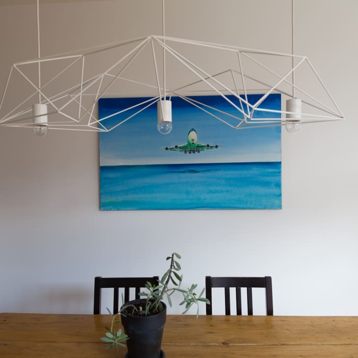 Dining room with wooden table, geometric chandelier, plant, and airplane painting on the wall.