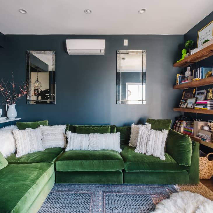 Green velvet sectional sofa with white pillows, floating wooden shelves with books and decor, and dark blue walls.