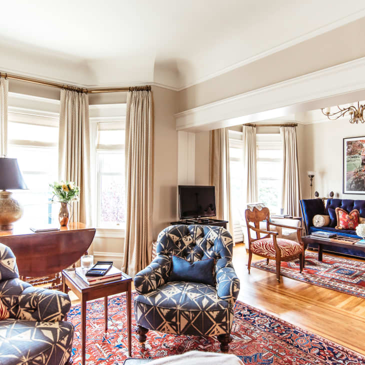 Elegant living room with patterned armchairs, blue velvet sofa, red and blue rugs, and large windows with beige curtains.