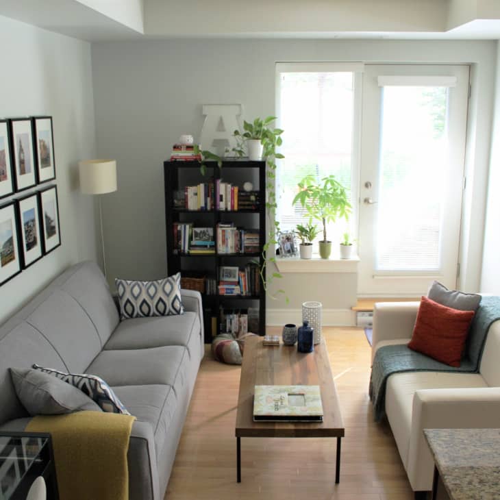 Living room with gray and white sofas, framed photos, bookshelf, and potted plants near a window.