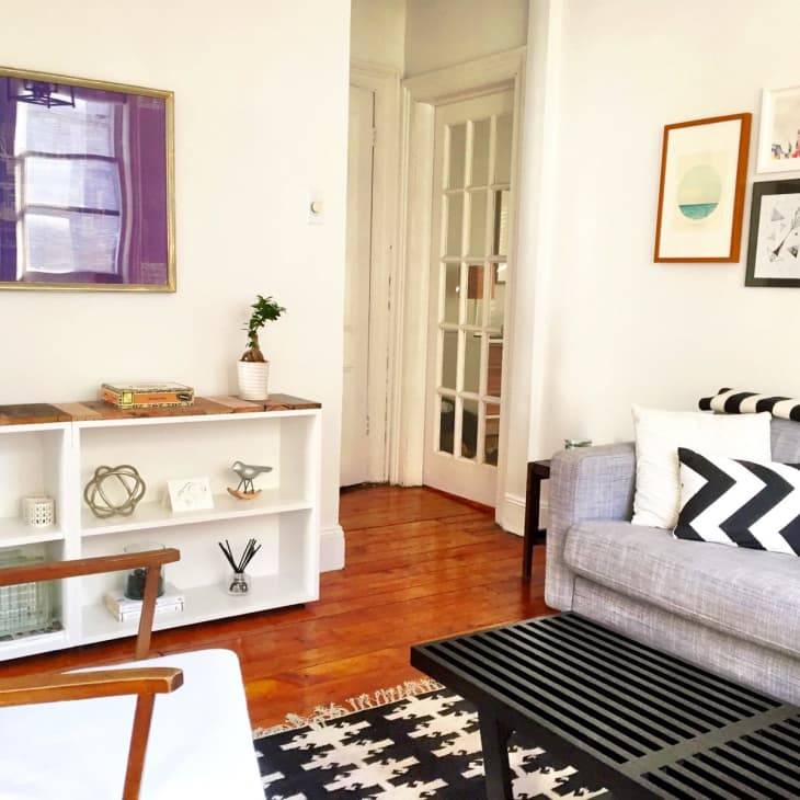Living room with gray sofa, black and white pillows, wooden chair, bookshelf, and framed art on white walls.
