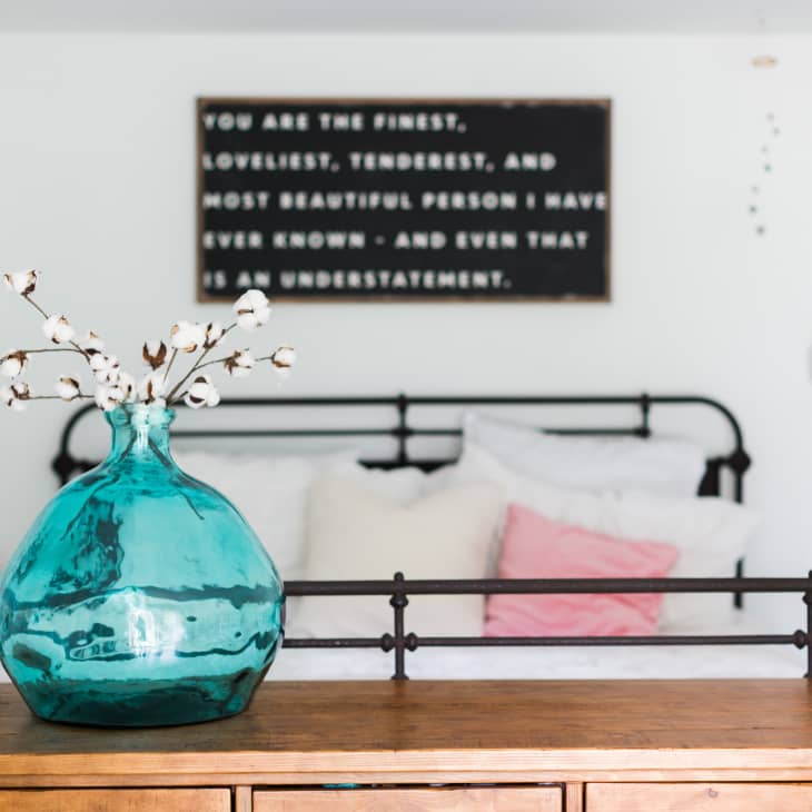 Bedroom with a metal bed frame, pink pillow, turquoise vase with branches, and a wall quote.