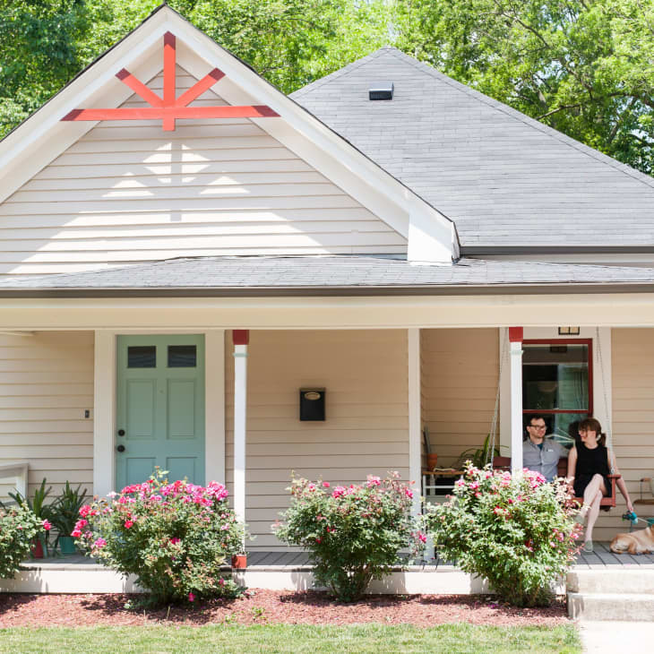 Front porch of a beige house with a blue door, red trim, two people sitting, and a dog lying nearby.