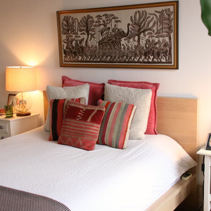 Cozy bedroom with a white bed, red and striped pillows, tribal wall art, and warm lighting from a bedside lamp.