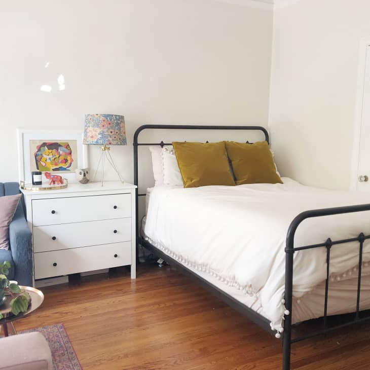 Bedroom with a black metal bed, mustard pillows, white dresser, floral lamp, and blue sofa on wooden floor.
