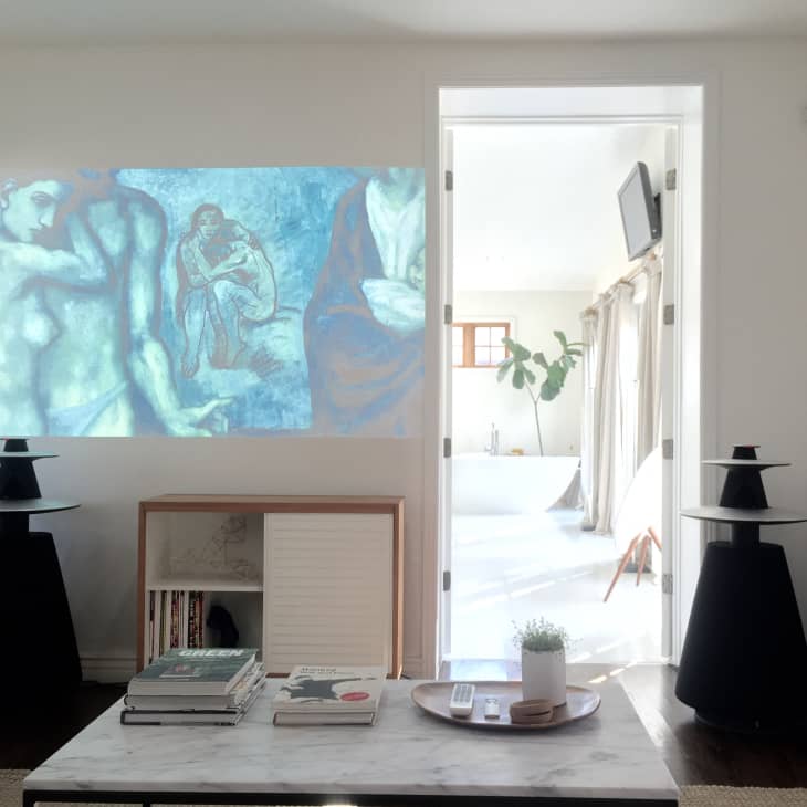 Living room with abstract art projection, black speakers, coffee table with books, and view into a bright bathroom.
