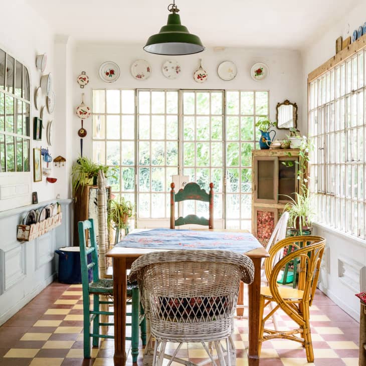 Eclectic dining room with mismatched chairs, large windows, decorative plates, and plants.