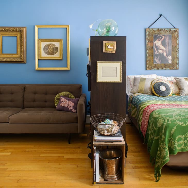 Brown sofa with floral pillows, blue accent wall, framed art, and a bed with green patterned bedding in a cozy room.
