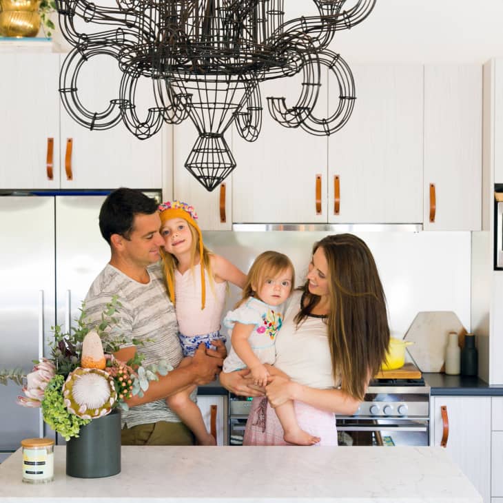 Family in a modern kitchen with white cabinets, black chandelier, and floral centerpiece on the counter.