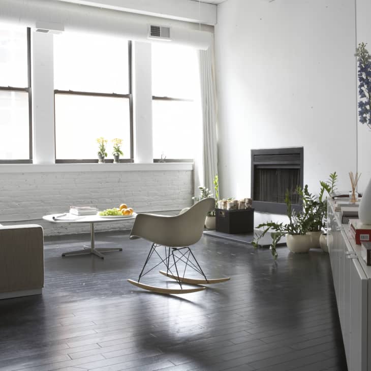 Modern living room with a beige sofa, white rocking chair, plants, and a fireplace, featuring large windows and dark wood flooring.