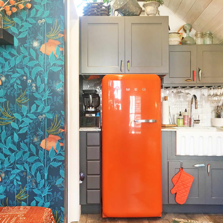 Colorful kitchen with orange Smeg fridge, gray cabinets, floral wallpaper, and farmhouse sink.