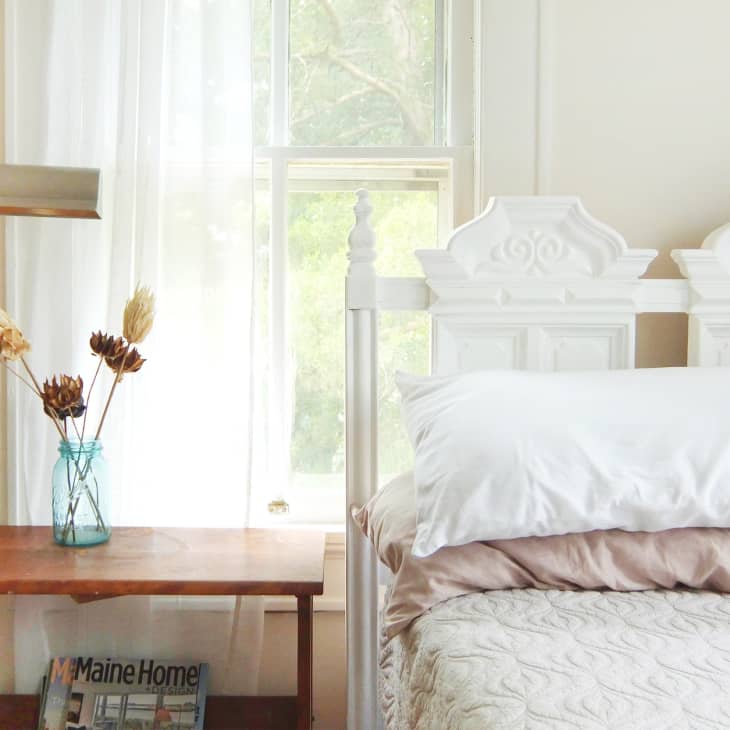 White ornate headboard bed with beige quilt, wooden side table, blue vase with dried flowers, and a brass lamp.