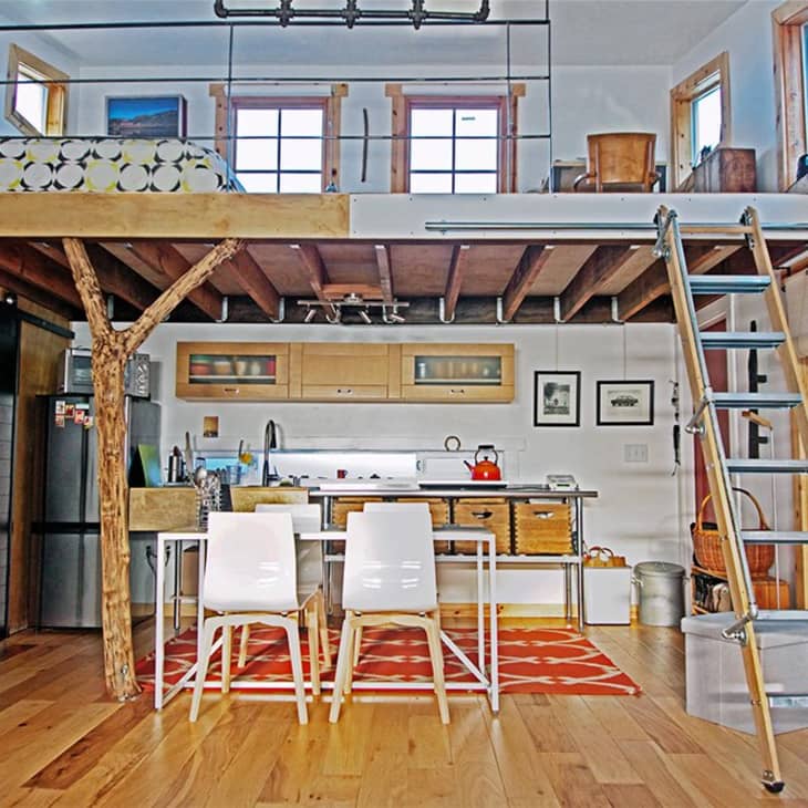 Loft apartment with wooden floors, a kitchen, dining table, ladder to bed, and rustic tree trunk support.