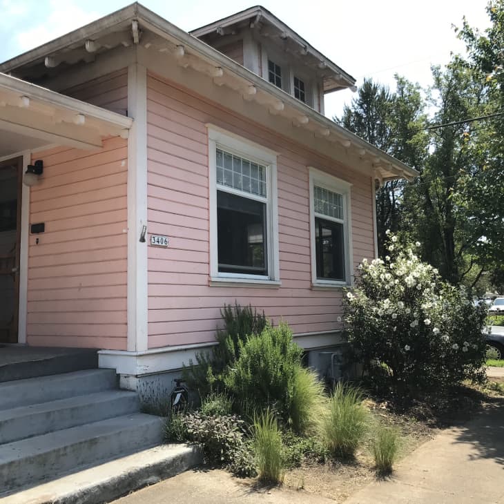 Pink house with white trim, front steps, and a garden with bushes and trees on a sunny day.