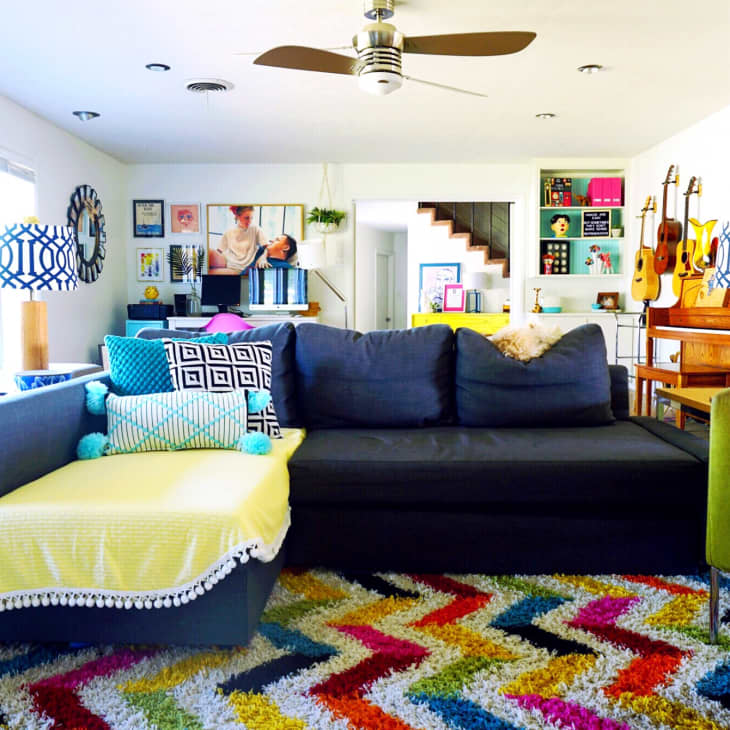 Colorful living room with a dark sofa, patterned pillows, vibrant rug, guitars, and eclectic wall art.