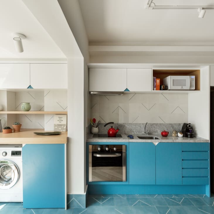 Modern kitchen with blue cabinets, stainless steel appliances, red kettle, and potted plants on wooden shelves.