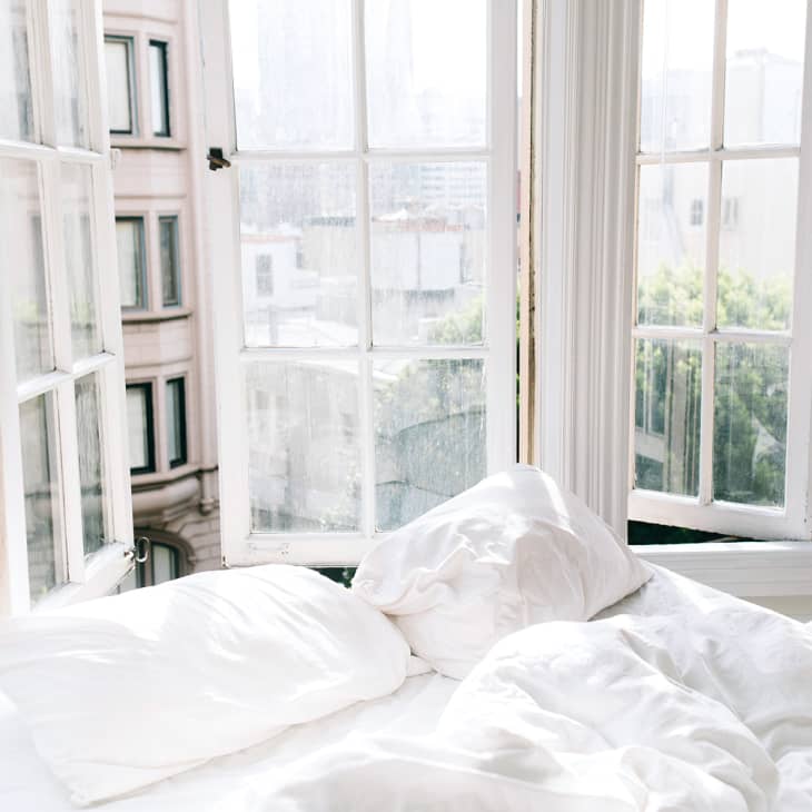 Unmade bed with white sheets in a bright room with large open windows overlooking city buildings.