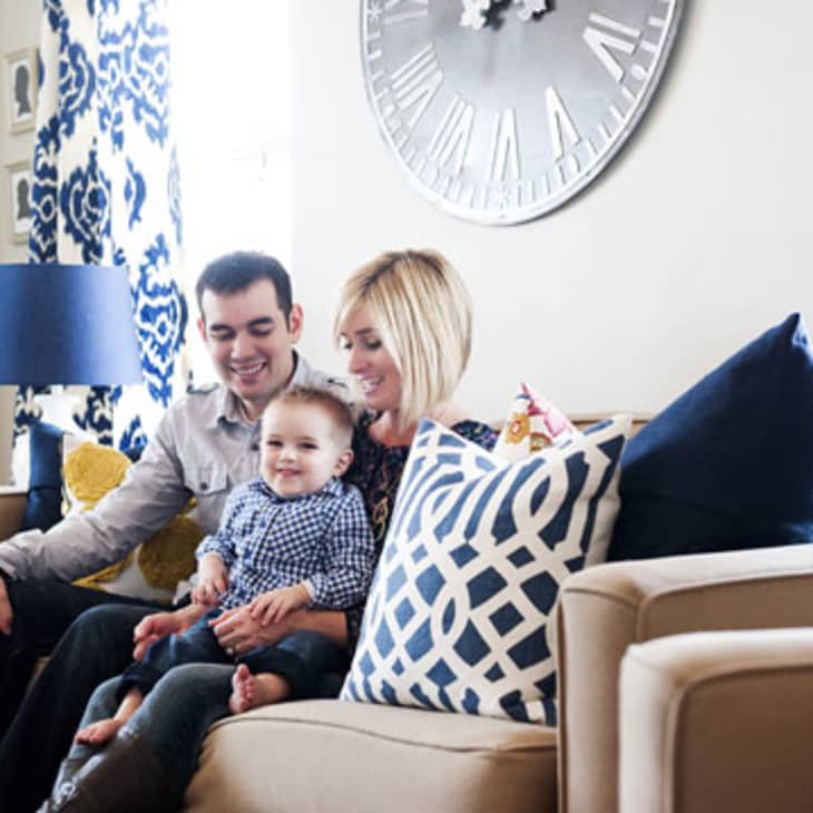 Family sitting on a beige sofa with blue and white patterned pillows, large clock on wall, and blue lamp in living room.