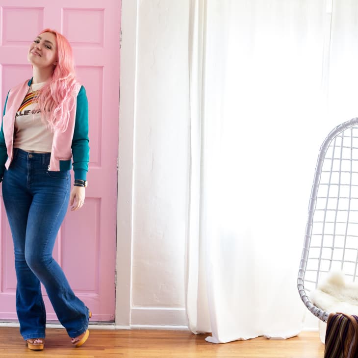 Woman with pink hair in casual outfit standing by a pink door, next to a hanging chair with a cushion.