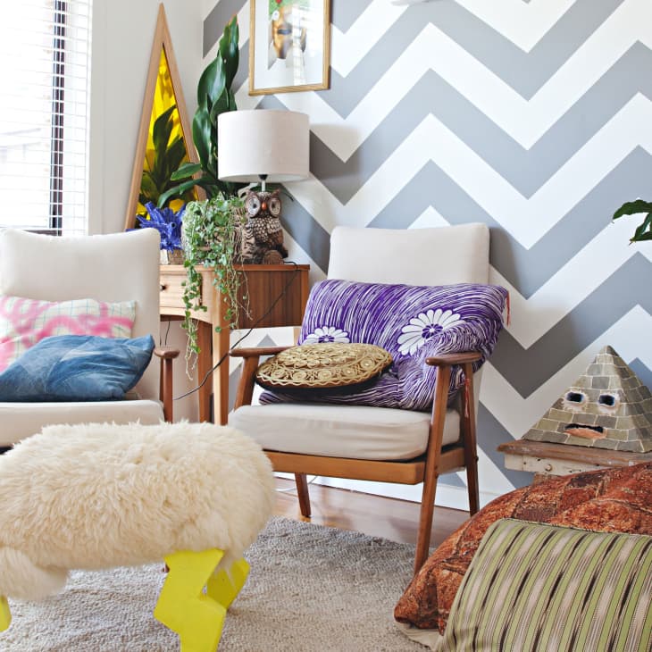 Chevron-patterned wall in a living room with white chairs, colorful cushions, plants, and eclectic decor.