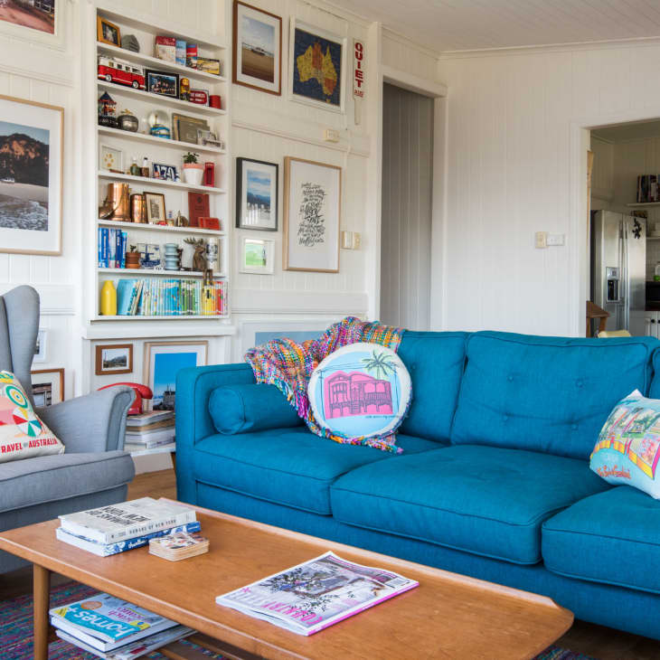 Living room with blue sofa, colorful pillows, gray armchair, and wall art; open view to kitchen with white cabinets.
