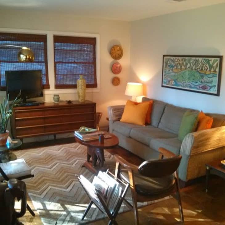 Cozy living room with a gray sofa, orange pillows, wooden furniture, plants, and decorative wall art.