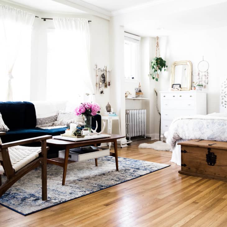 Bright bedroom with a cozy seating area, wooden floors, floral bedspread, wooden chest, and pink flowers on a coffee table.