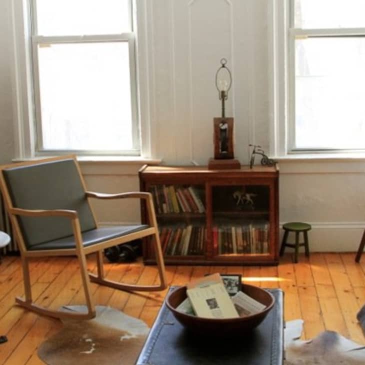 Cozy living room with wooden floors, rocking chair, bookshelf, and a small table with a lamp between two windows.