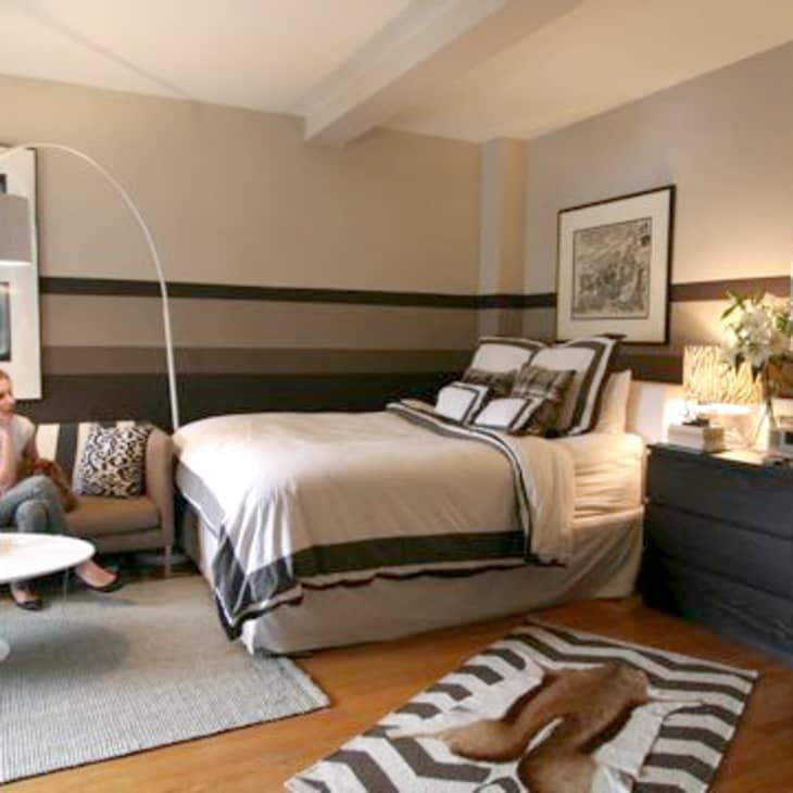 Cozy bedroom with striped walls, bed, sofa, round table, and dresser with flowers.