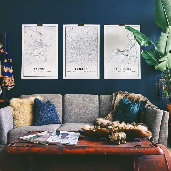 Gray sofa with blue and yellow pillows, wooden coffee table, large plant, and three city map posters on a dark blue wall.