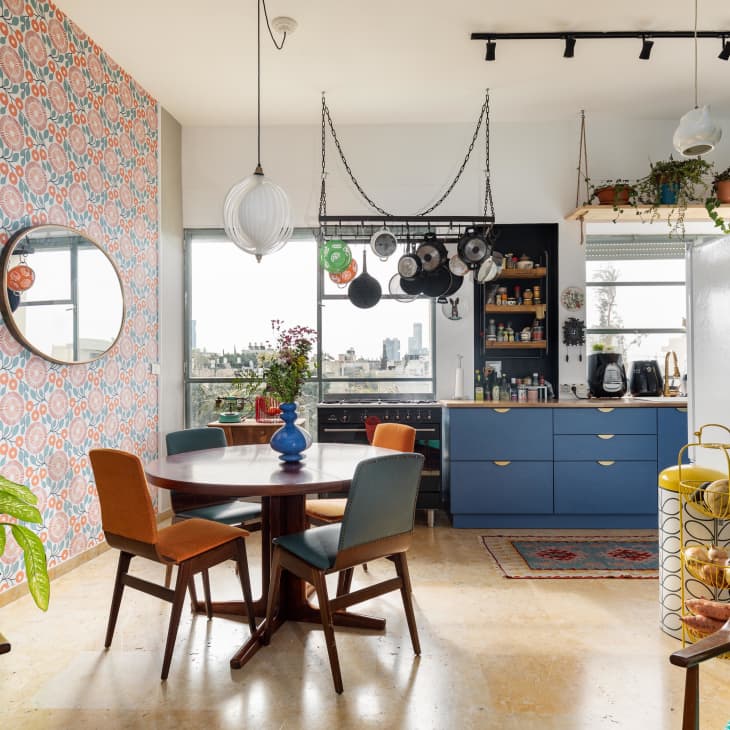Eclectic kitchen and dining area with patterned wallpaper, round table, blue cabinets, hanging pots, and plants.