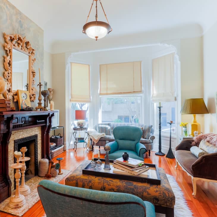 Eclectic living room with vintage fireplace, ornate mirror, teal armchair, and large windows with beige blinds.