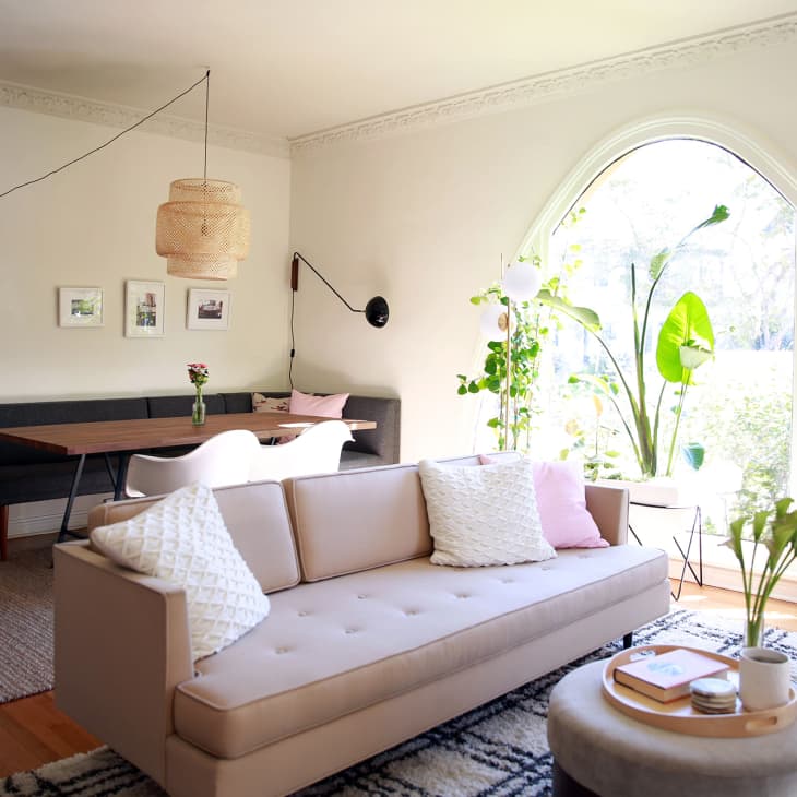 Living room with beige sofa, round ottoman, wooden dining table, black bench, and large arched window with plants.