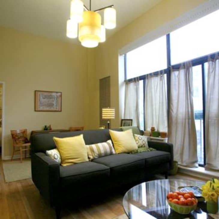 Living room with dark sofa, yellow pillows, large windows, and a round glass coffee table with fruit and flowers.