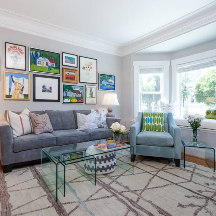 Living room with gray sofa, blue armchair, glass coffee table, and gallery wall. Large windows with green cushions on bench.