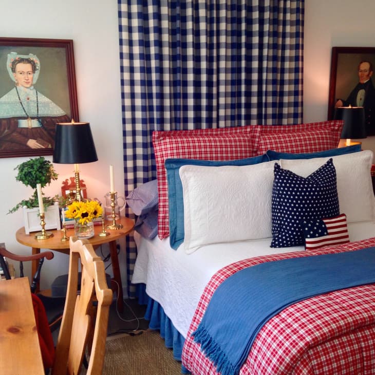 Cozy bedroom with red plaid bedding, blue checkered curtains, and patriotic pillows. Vintage portraits and sunflowers on side table.