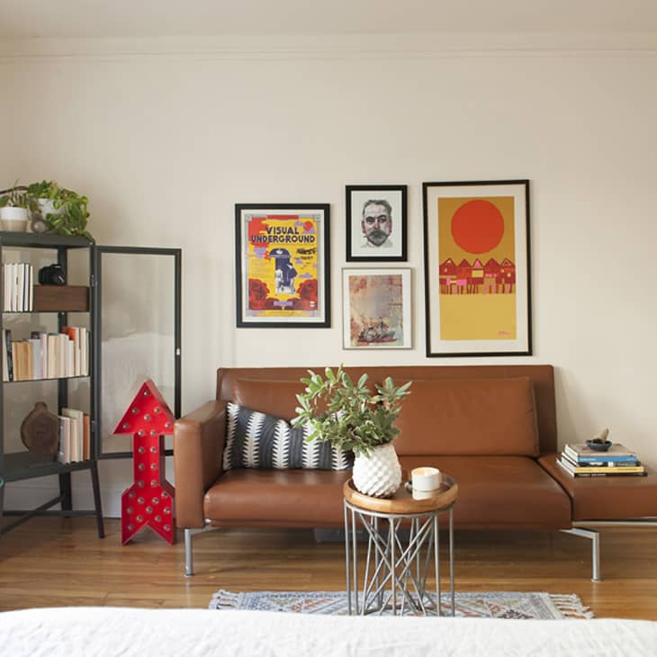 Living room with brown leather sofa, framed art, glass cabinet, plants, and a small round table with books and decor.