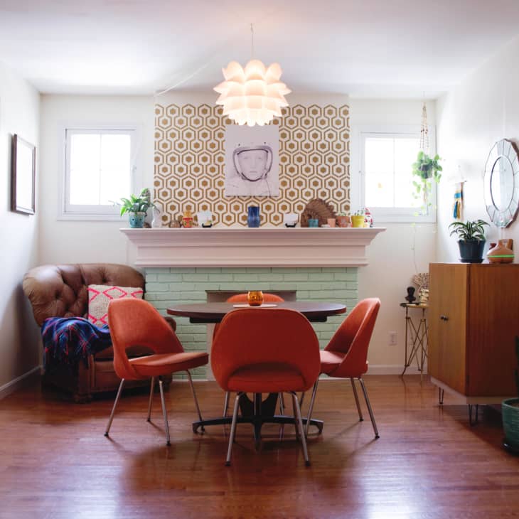 Mid-century modern dining room with orange chairs, round table, patterned wallpaper, and plants on a mint green fireplace mantel.