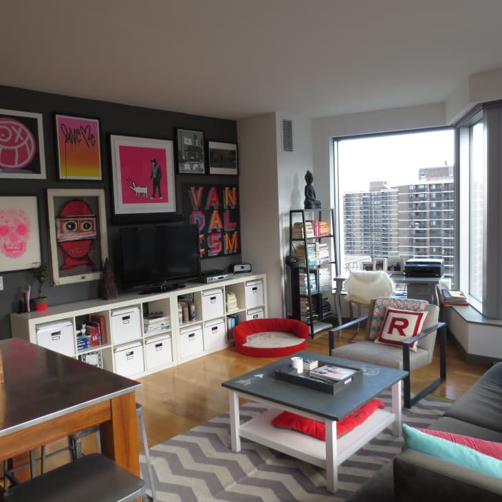 Modern living room with colorful art wall, gray sofa, chevron rug, and large windows overlooking cityscape.