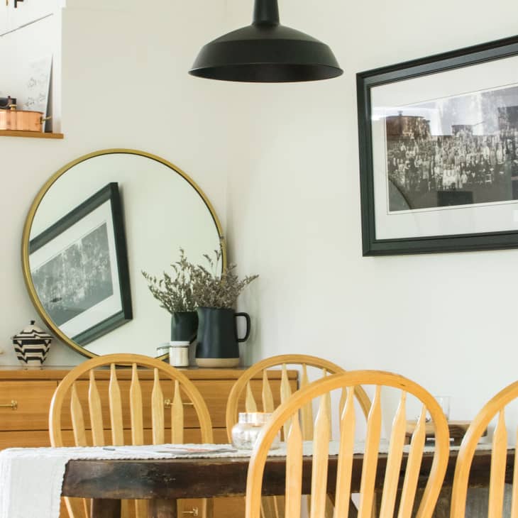 Dining area with wooden chairs, round table, black pendant light, mirror, framed photo, and dresser with books and decor.