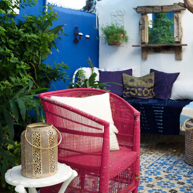 Colorful patio with pink chair, blue and white tiles, wicker furniture, and lush greenery against a blue wall.