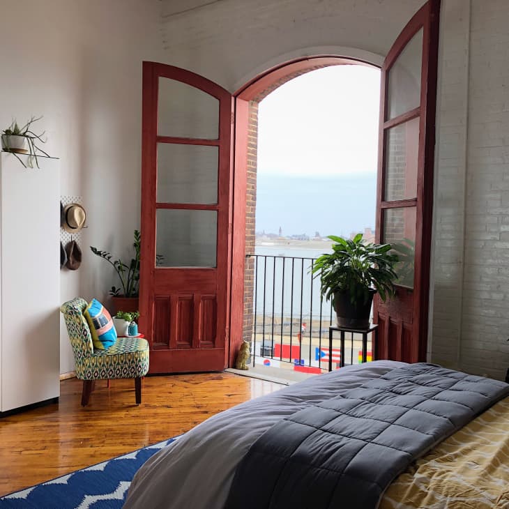 Bedroom with open red doors to a balcony, overlooking water. Features a patterned chair, potted plants, and a blue rug.