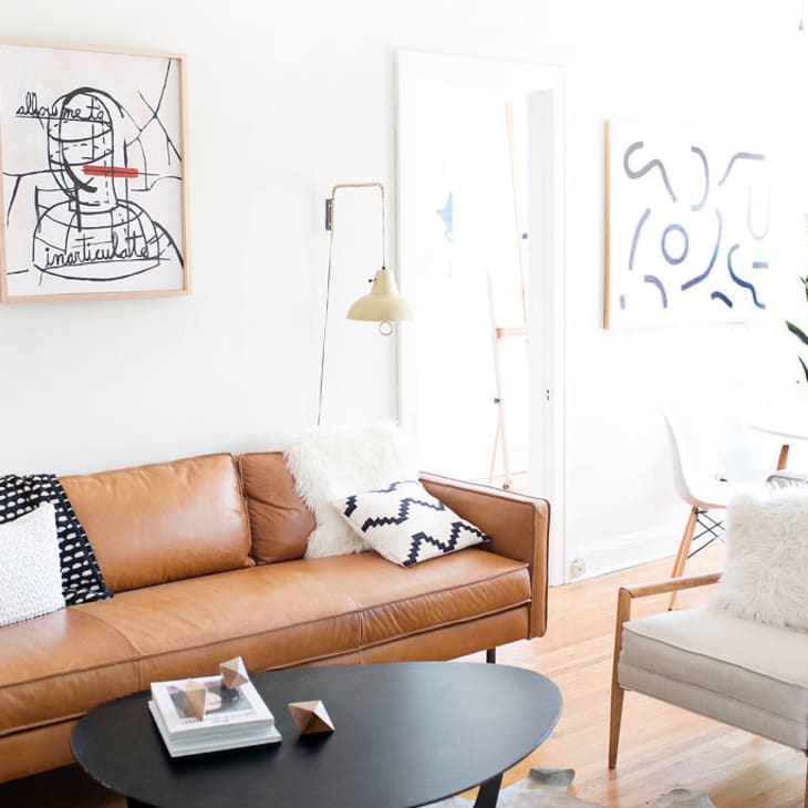 Mid-century modern living room with tan leather sofa, abstract art, black coffee table, and dining area with a potted plant.