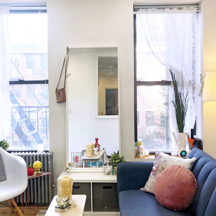 Cozy living room with blue sofa, white chair, floor lamp, and window with sheer curtains. Decor includes plants and art.