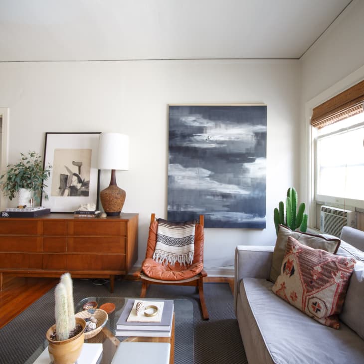 Living room with a gray sofa, patterned pillows, wooden dresser, abstract painting, and potted plants.