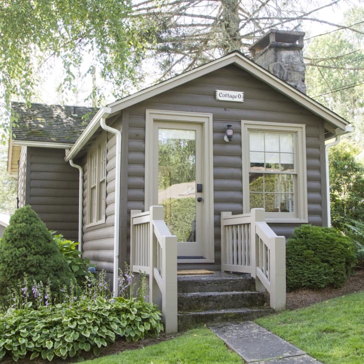 Small brown log cabin with a stone chimney, surrounded by lush greenery and a wooden porch.